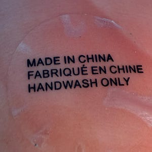 Puede incluir: Primer plano de una superficie color melocot&oacute;n con una pegatina transparente. La pegatina tiene texto negro que dice: "MADE IN CHINA", "FABRIQU&Eacute; EN CHINE" y "HANDWASH ONLY".