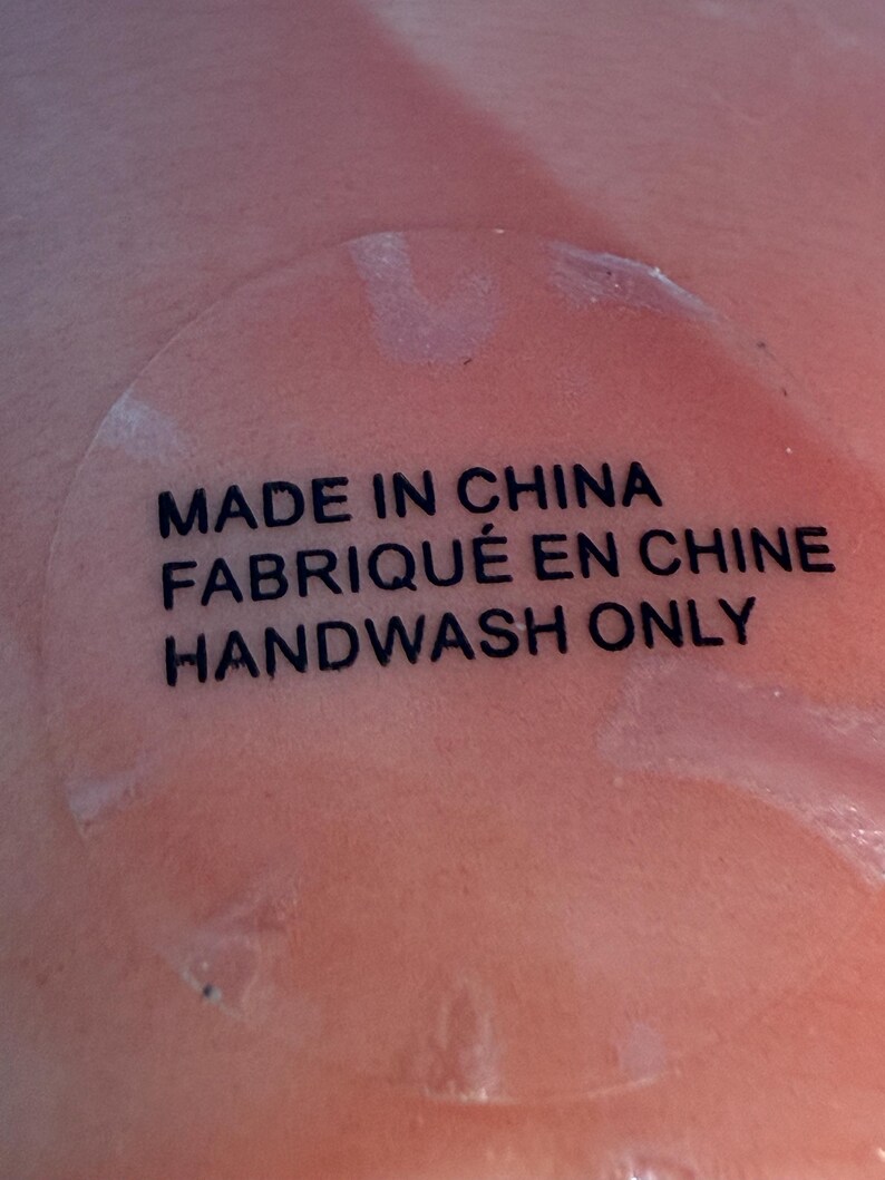 Puede incluir: Primer plano de una superficie color melocot&oacute;n con una pegatina transparente. La pegatina tiene texto negro que dice: "MADE IN CHINA", "FABRIQU&Eacute; EN CHINE" y "HANDWASH ONLY".