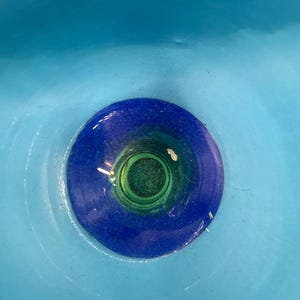 May include: A close-up view of a glass object with a vibrant color scheme. The piece features a central green core, encircled by a deep blue ring, and set against a light blue backdrop. The glass appears smooth and reflective.