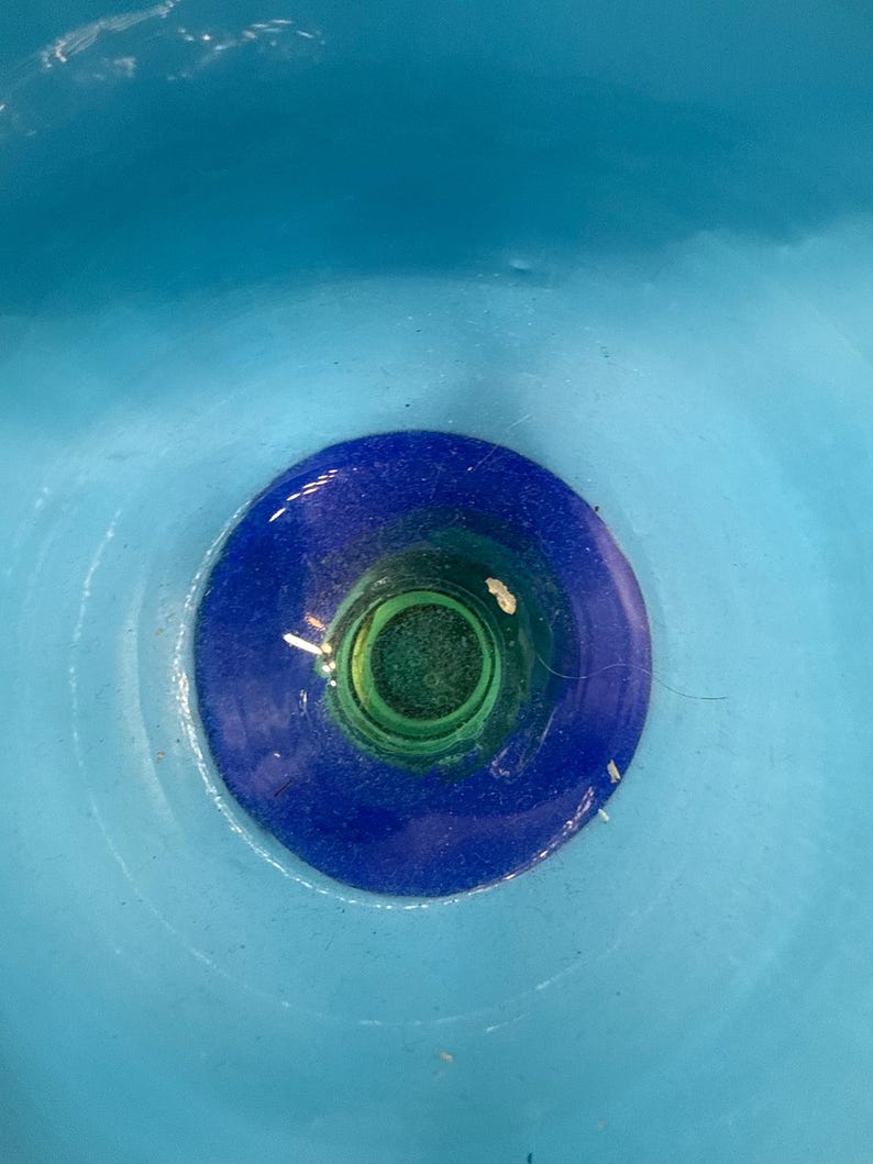 May include: A close-up view of a glass object with a vibrant color scheme. The piece features a central green core, encircled by a deep blue ring, and set against a light blue backdrop. The glass appears smooth and reflective.