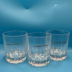 May include: Three clear, short whiskey glasses with the "Johnnie Walker" logo printed in gold. The glasses have a textured, vertical design and are set against a blue background. Ideal for serving spirits.