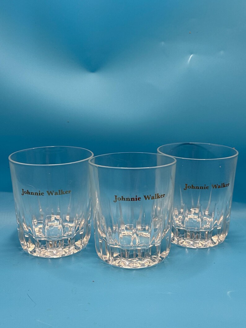 May include: Three clear, short whiskey glasses with the "Johnnie Walker" logo printed in gold. The glasses have a textured, vertical design and are set against a blue background. Ideal for serving spirits.