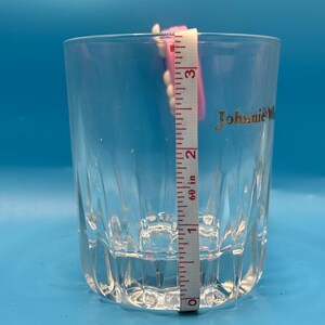 May include: A clear, faceted glass with the gold text "Johnnie Walker" on the side. A pink and white measuring tape is placed inside the glass, showing a height of approximately 3 inches.