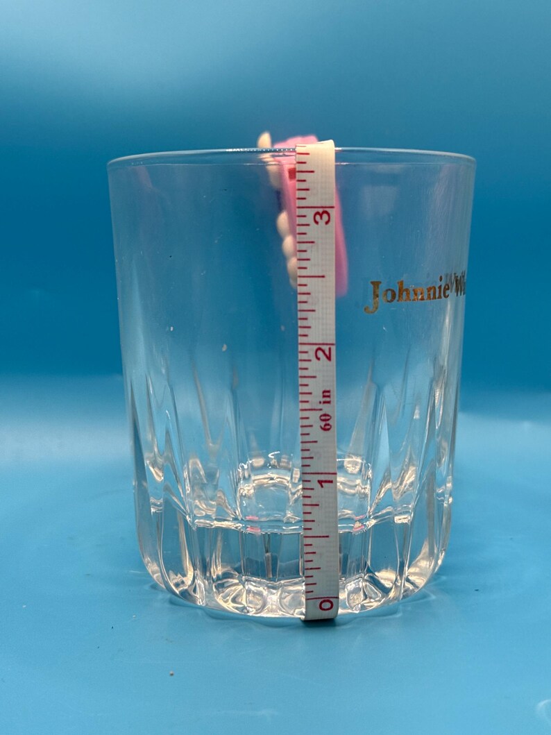 May include: A clear, faceted glass with the gold text "Johnnie Walker" on the side. A pink and white measuring tape is placed inside the glass, showing a height of approximately 3 inches.
