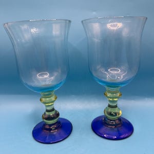 May include: Two clear glass goblets with a blue rim and base, and a yellow stem. The goblets have a tulip-shaped bowl and are set against a light blue background. The base is approximately 3 inches in diameter.