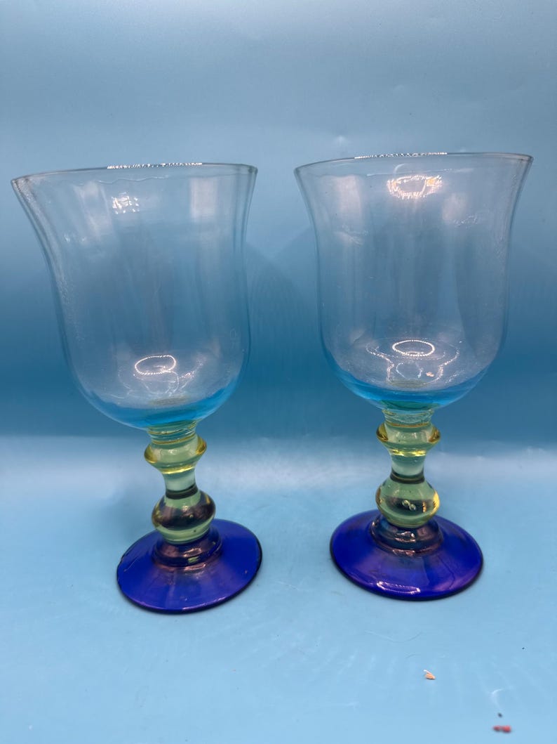 May include: Two clear glass goblets with a blue rim and base, and a yellow stem. The goblets have a tulip-shaped bowl and are set against a light blue background. The base is approximately 3 inches in diameter.