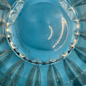 May include: Close-up of a clear, turquoise-colored glass bowl. The bowl has a scalloped edge and a circular design in the center. The glass reflects light, creating highlights and shadows.