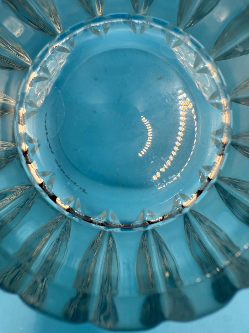 May include: Close-up of a clear, turquoise-colored glass bowl. The bowl has a scalloped edge and a circular design in the center. The glass reflects light, creating highlights and shadows.
