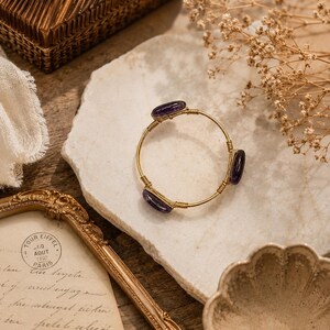 May include: A gold-toned bangle bracelet with three oval, dark purple gemstones. The bracelet is set on a white stone surface, with a vintage-style frame and a small woven basket in the background. The bracelet's design is simple and elegant.