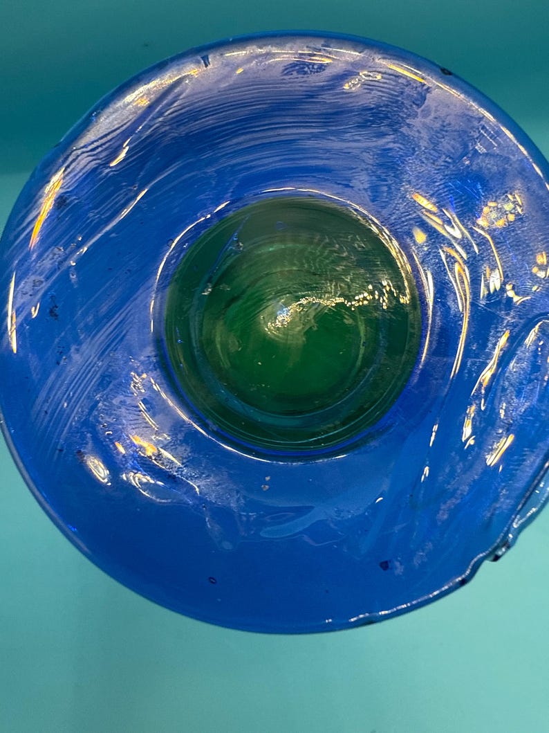 May include: A handcrafted glass bowl with a vibrant blue exterior and a green interior. The bowl's design features a circular shape with a smooth, glossy finish. The glass appears to have a textured, rippled effect, adding visual interest to the piece.