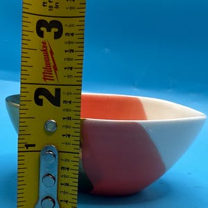 Puede incluir: Un peque&ntilde;o cuenco de cer&aacute;mica colorido con un dise&ntilde;o geom&eacute;trico en rojo, blanco y verde. El cuenco mide aproximadamente 6,3 cm de ancho y 3,8 cm de alto. El cuenco est&aacute; sobre un fondo azul.