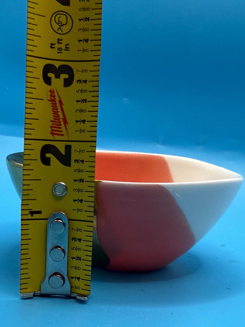 Puede incluir: Un peque&ntilde;o cuenco de cer&aacute;mica colorido con un dise&ntilde;o geom&eacute;trico en rojo, blanco y verde. El cuenco mide aproximadamente 6,3 cm de ancho y 3,8 cm de alto. El cuenco est&aacute; sobre un fondo azul.