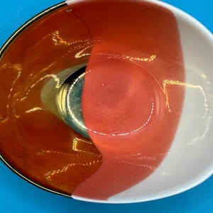 Puede incluir: Un cuenco decorativo peque&ntilde;o con borde dorado e interior dividido. Un lado es naranja vibrante, el otro es blanco con una franja color coral. Una pieza de metal plateado est&aacute; en el centro del cuenco.