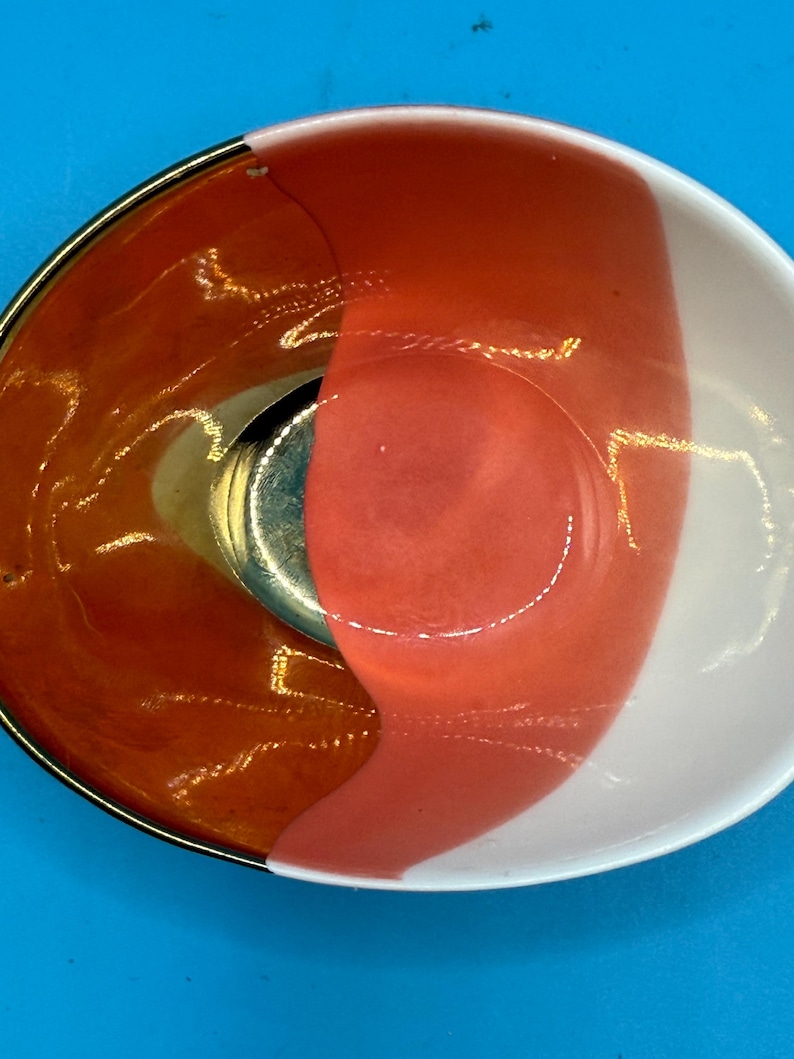 Puede incluir: Un cuenco decorativo peque&ntilde;o con borde dorado e interior dividido. Un lado es naranja vibrante, el otro es blanco con una franja color coral. Una pieza de metal plateado est&aacute; en el centro del cuenco.