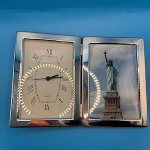 May include: A silver-colored hinged frame with a clock on one side and a picture of the Statue of Liberty on the other. The clock face has Roman numerals and the word "QUARTZ". The picture frame has the words "Thank You" at the top.