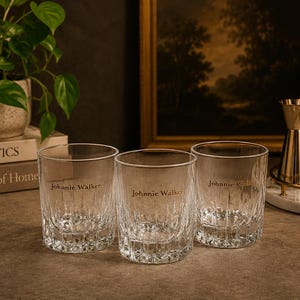 May include: Three clear, textured whiskey glasses with the "Johnnie Walker" logo etched on the front. The glasses are arranged on a dark surface, with a gold cocktail shaker and other bar accessories in the background. A potted plant and books are also visible.