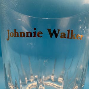 May include: Clear, faceted glass tumbler with the gold text "Johnnie Walker" visible. The glass has a classic design, suitable for serving whiskey or other beverages. The background is a light blue.