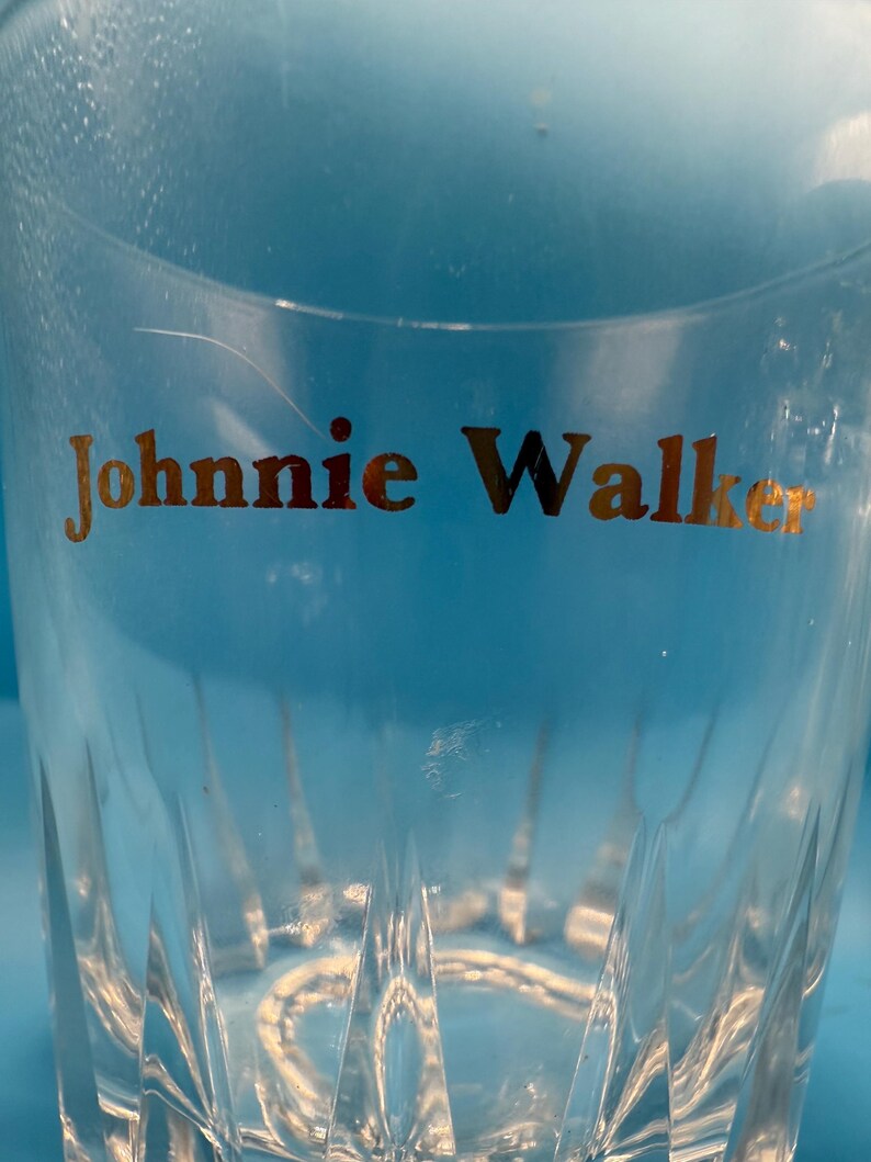 May include: Clear, faceted glass tumbler with the gold text "Johnnie Walker" visible. The glass has a classic design, suitable for serving whiskey or other beverages. The background is a light blue.