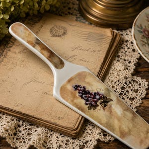 May include: A white and brown ceramic cake server with a grape cluster design. The server rests on a stack of vintage letters and a lace doily, with a brass candlestick and floral arrangement in the background.