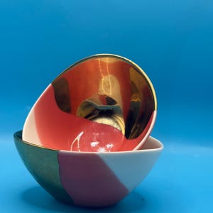 Puede incluir: Dos cuencos decorativos de cer&aacute;mica. El cuenco superior tiene un interior dorado y un exterior rojo y blanco. El cuenco inferior tiene un exterior blanco, rosa y verde. Los cuencos est&aacute;n apilados uno encima del otro sobre un fondo azul.