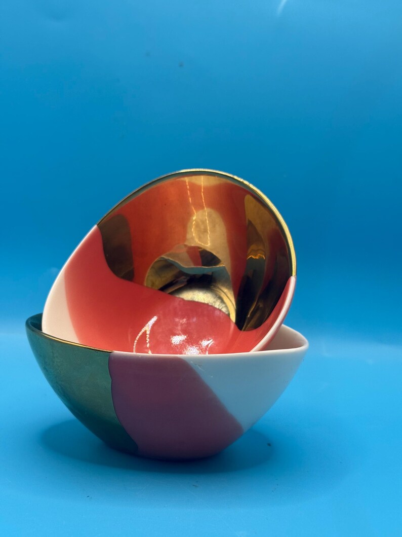 Puede incluir: Dos cuencos decorativos de cer&aacute;mica. El cuenco superior tiene un interior dorado y un exterior rojo y blanco. El cuenco inferior tiene un exterior blanco, rosa y verde. Los cuencos est&aacute;n apilados uno encima del otro sobre un fondo azul.