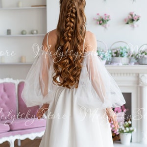 Puede incluir: Una mujer con largo cabello castaño rojizo peinado en una trenza, vistiendo un vestido blanco con mangas abullonadas transparentes. El vestido tiene un diseño simple y elegante. El fondo incluye una chimenea blanca, un sofá rosa y decoraciones florales.