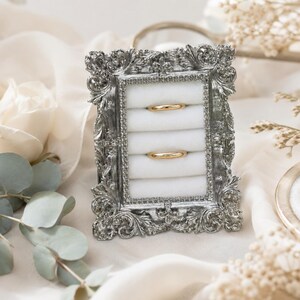 May include: A rectangular ring display stand featuring an ornate silver-toned frame with intricate scrollwork and a beaded inner border. The center contains five horizontal white velvet slots, with two plain gold bands resting in the top and fourth slots.