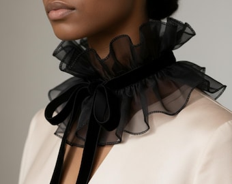 May include: Black ruffled collar made of sheer fabric and velvet. The collar has a high, ruffled neckline and a velvet band that ties in a bow at the front. The collar is worn over a light-colored garment.