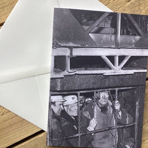 May include: Black and white photograph of three men wearing hard hats in a mine shaft. The men are standing in a cage, looking out at the camera. The cage is surrounded by metal bars and chains.