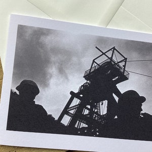 May include: Black and white photograph of two silhouetted figures wearing hard hats standing in front of a large industrial structure. The structure is a mine shaft headframe with a large pulley system and cables.