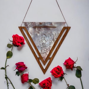 Puede incluir: Un colgante de pared triangular de madera con un diseño floral y una cadena plateada. El triángulo está hecho de dos piezas de madera, una encima de la otra. El diseño floral está impreso sobre un fondo blanco. La cadena está unida a la parte superior del triángulo. Hay rosas rojas alrededor del triángulo.