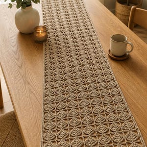 Könnte beinhalten: Beige gehäkelter Tischläufer mit sich wiederholendem Blattmuster. Der Läufer liegt auf einem Holztisch, mit einer Vase mit Grünzeug, einer Kerze und einer Tasse. Der Läufer verleiht dem Essbereich einen Hauch von Handwerkskunst.