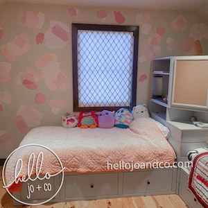 Op de afbeelding: Een slaapkamer met een lichtroze en witte gepatroneerde muur, een wit bed met een roze en witte deken en een witte boekenkast met een kurkdeur. Het bed heeft een paar knuffels erop en de tekst "hello jo + co" staat op het bed.