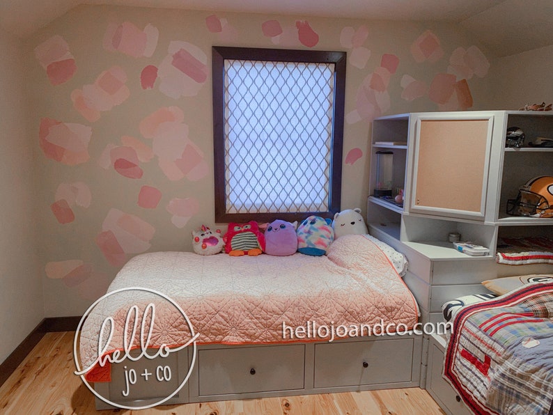 Op de afbeelding: Een slaapkamer met een lichtroze en witte gepatroneerde muur, een wit bed met een roze en witte deken en een witte boekenkast met een kurkdeur. Het bed heeft een paar knuffels erop en de tekst "hello jo + co" staat op het bed.