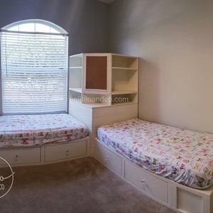 Op de afbeelding: Twee eenpersoonsbedden met witte frames en lades, elk met een wit en roze beddengoed. De bedden staan in een hoek van een kamer met een witte ingebouwde plankeneenheid met een kurk achterwand. De plankeneenheid heeft drie planken en een lade. De kamer heeft witte muren en een raam met witte jaloezie&euml;n.