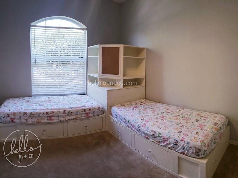 Op de afbeelding: Twee eenpersoonsbedden met witte frames en lades, elk met een wit en roze beddengoed. De bedden staan in een hoek van een kamer met een witte ingebouwde plankeneenheid met een kurk achterwand. De plankeneenheid heeft drie planken en een lade. De kamer heeft witte muren en een raam met witte jaloezie&euml;n.