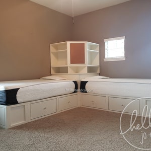 Op de afbeelding: Witte hoekbedframe met ingebouwde planken en laden. Het bedframe heeft twee eenpersoonsmatrassen en een wit hoofdbord met een kurkboordeinsatz. Het bedframe staat in een kamer met beige muren en twee ramen.