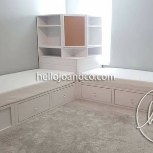 Op de afbeelding: Twee witte eenpersoonsbedden met opbergladen en een witte hoekkast met een kurk achterwand. De bedden staan in een hoek van een kamer met witte muren en een tapijt vloer. De tekst "hellojoandco.com" is zichtbaar op de bedden. De tekst "hello jo + co" is zichtbaar op de vloer.
