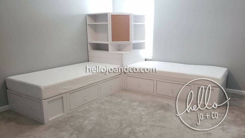 Op de afbeelding: Twee witte eenpersoonsbedden met opbergladen en een witte hoekkast met een kurk achterwand. De bedden staan in een hoek van een kamer met witte muren en een tapijt vloer. De tekst "hellojoandco.com" is zichtbaar op de bedden. De tekst "hello jo + co" is zichtbaar op de vloer.