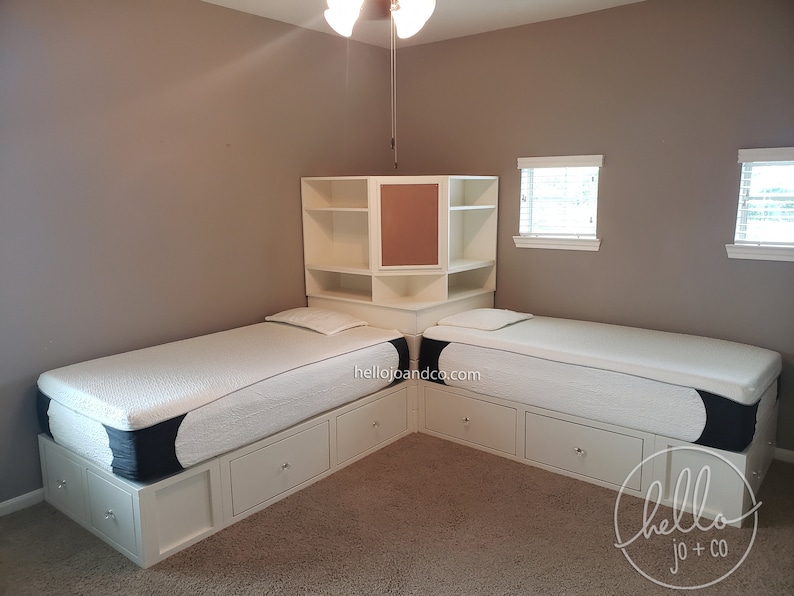 Op de afbeelding: Twee eenpersoonsbedden met witte houten frames en laden, geplaatst in een hoek van een kamer met beige muren. Een witte houten plankeneenheid met een bruin achterpaneel is boven de bedden gemonteerd. De bedden hebben witte matrassen met zwart-witte patroonhoezen. De kamer heeft een beige tapijt en twee ramen met witte jaloezie&euml;n.