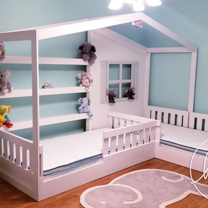 May include: White wooden house-shaped bed frame with a built-in bookshelf and a window cutout. The bed has a white mattress and a white railing. There are stuffed animals on the shelves and a gray rug on the floor. The text "hello jo + co" is visible on the rug.