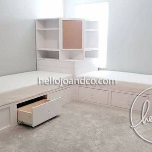 Op de afbeelding: Twee witte eenpersoonsbedden met opbergladen en een witte hoekkast met een bruin achterpaneel. De bedden staan in een hoek van een kamer met witte muren en een tapijt. De tekst "hellojoandco.com" is zichtbaar op de bedden.