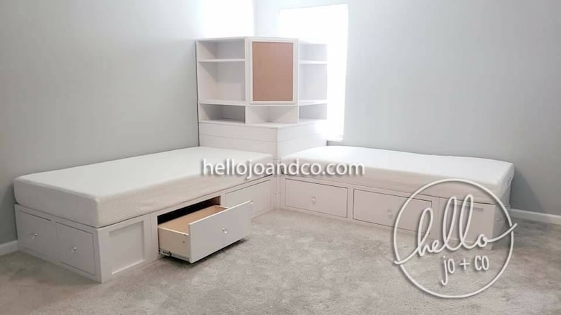 Op de afbeelding: Twee witte eenpersoonsbedden met opbergladen en een witte hoekkast met een bruin achterpaneel. De bedden staan in een hoek van een kamer met witte muren en een tapijt. De tekst "hellojoandco.com" is zichtbaar op de bedden.