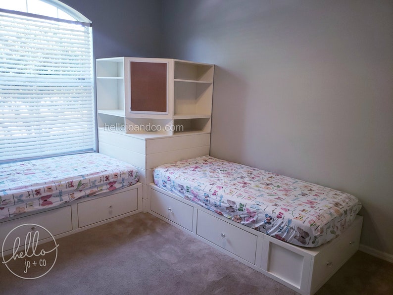 Op de afbeelding: Twee eenpersoonsbedden met witte frames en lades, elk met een bedsprei met patroon. De bedden staan in een hoek met een witte ingebouwde plankeneenheid met een bruine kurkplankdeur. De tekst "hellojoandco.com" is zichtbaar op de plankeneenheid.