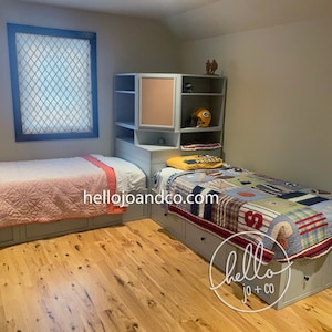 Op de afbeelding: Twee eenpersoonsbedden met opbergladen, een met een roze en witte deken en de andere met een kleurrijk patchwork-dekbed. De bedden staan in een kamer met lichtgrijze muren en houten vloer. Een hoekkast met een prikbord bevindt zich achter het bed met het patchwork-dekbed. De kast heeft een paar decoratieve items, waaronder een voetbalhelm.