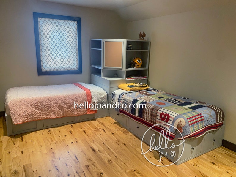 Op de afbeelding: Twee eenpersoonsbedden met opbergladen, een met een roze en witte deken en de andere met een kleurrijk patchwork-dekbed. De bedden staan in een kamer met lichtgrijze muren en houten vloer. Een hoekkast met een prikbord bevindt zich achter het bed met het patchwork-dekbed. De kast heeft een paar decoratieve items, waaronder een voetbalhelm.