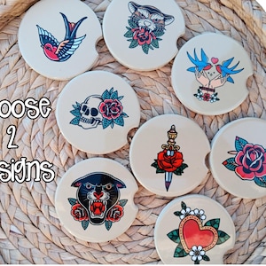May include: Eight round ceramic car coasters with a notch on the side, featuring colorful traditional tattoo-style illustrations including a swallow, panther, skull, rose, dagger, and heart. The text Choose 2 designs is printed on the left side.