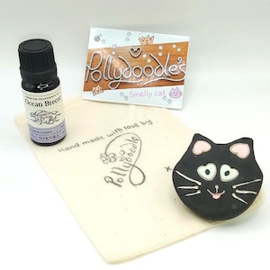 May include: A small, black, glass bottle of essential oil with a white label that says "Ocean Breeze" and a white muslin bag with a black cat face charm and the text "Hand made with love by Pollydoodle" printed on it.