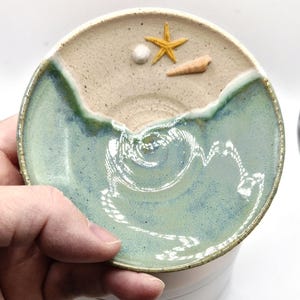 May include: A ceramic dish depicting a beach scene. The top half is sandy beige with a yellow starfish, a white shell, and a brown seashell. The bottom half is light blue, representing the ocean, with a wave design.