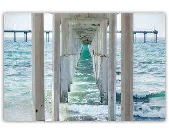 May include: A view through the concrete pillars of a pier, looking out to the ocean. The water is a vibrant turquoise blue, with white waves crashing against the sand.